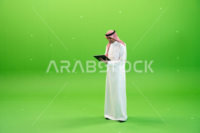 البحث عن عمل عن طريق الانترنت، كروما لرجل عربي خليجي يرتدي الزي التقليدي السعودي ويحمل بيده جهاز التابليت، التسويق الإلكتروني عبر الأجهزة الحديثة، تصفح مواقع التواصل الإجتماعي، متابعه آخر أخبار السوشال ميديا، صورة الجسم بالطول الكامل، خلفية خضراء