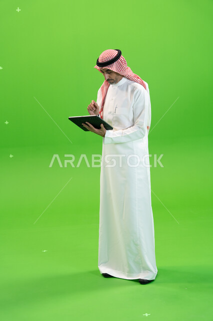 البحث عن عمل عن طريق الانترنت، كروما لرجل عربي خليجي يرتدي الزي التقليدي السعودي ويحمل بيده جهاز التابليت، التسويق الإلكتروني عبر الأجهزة الحديثة، تصفح مواقع التواصل الإجتماعي، متابعه آخر أخبار السوشال ميديا، صورة الجسم بالطول الكامل، خلفية خضراء