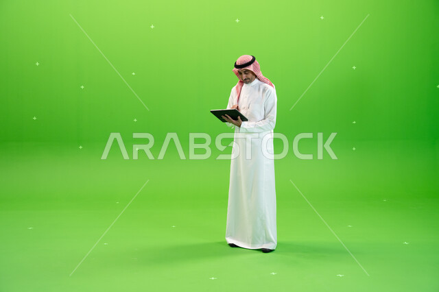 البحث عن عمل عن طريق الانترنت، كروما لرجل عربي خليجي يرتدي الزي التقليدي السعودي ويحمل بيده جهاز التابليت، التسويق الإلكتروني عبر الأجهزة الحديثة، تصفح مواقع التواصل الإجتماعي، متابعه آخر أخبار السوشال ميديا، صورة الجسم بالطول الكامل، خلفية خضراء