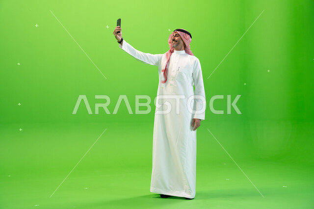 التقاط صور السيلفي التذكارية، إجراء مكالمة فيديو عبر الجوال، كروما لرجل عربي خليجي سعودي يرتدي الثوب والشماغ السعودي يقوم بإجراء مكالمة فيديو عبر الهاتف المحمول، التواصل مع الأصدقاء والأقارب بإيماءات السعادة، صورة الجسم بالطول الكامل، خلفية خضراء