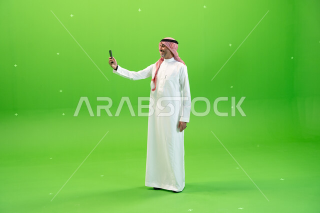 Taking memorial selfies, making a video call via mobile phone, Chroma of a Saudi Arabian Gulf man wearing the Saudi thobe and shemagh making a video call via mobile phone, communicating with friends and relatives with gestures of happiness, full-length bo