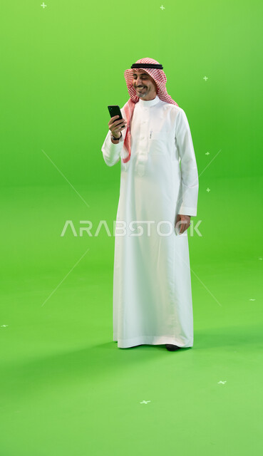 إجراء مكالمة فيديو عبر الجوال، تبادل النقاشات والأحاديث، كروما لرجل عربي خليجي سعودي يرتدي الثوب والشماغ السعودي يقوم بمتابعه أخبار السوشال ميديا عبر الهاتف المحمول، التواصل مع الأصدقاء والأقارب بإيماءات السعادة، صورة الجسم بالطول الكامل، خلفية خضراء