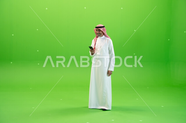 إجراء مكالمة فيديو عبر الجوال، تبادل النقاشات والأحاديث، كروما لرجل عربي خليجي سعودي يرتدي الثوب والشماغ السعودي يقوم بمتابعه أخبار السوشال ميديا عبر الهاتف المحمول، التواصل مع الأصدقاء والأقارب بإيماءات السعادة، صورة الجسم بالطول الكامل، خلفية خضراء