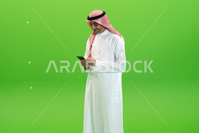 إرسال رسائل نصيه عبر الجوال، مفهوم صلة الرحم، كروما لرجل عربي خليجي سعودي يرتدي الثوب والشماغ السعودي يقوم بمتابعه أخبار السوشال ميديا عبر الهاتف المحمول، التواصل مع الأصدقاء والأقارب بإيماءات السعادة،  خلفية خضراء
