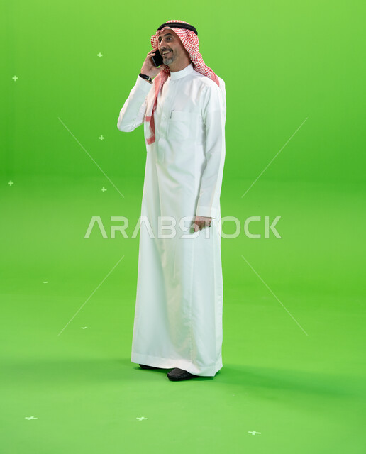 الاتفاق على موعد عمل، كروما لرجل عربي خليجي سعودي يرتدي الثوب والشماغ السعودي يقوم بإجراء مكالمة صوتيه عبر الهاتف المحمول، التواصل مع الأصدقاء والأقارب بإيماءات السعادة، مفهوم صلة الرحم، صورة الجسم بالطول الكامل، خلفية خضراء