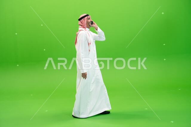 صورة الجسم بالطول الكامل ،التسامر والضحك  مع الأصدقاء والأقارب بإيماءات السعادة، كروما لرجل عربي خليجي سعودي يرتدي الثوب والشماغ السعودي يقوم بالتنقل في المكان أثناء إجراء مكالمة صوتيه عبر الهاتف المحمول، مفهوم صلة الرحم، خلفية خضراء