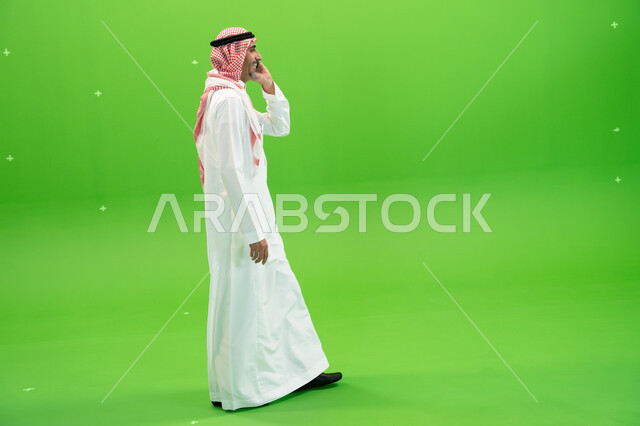 صورة الجسم بالطول الكامل ،التسامر والضحك  مع الأصدقاء والأقارب بإيماءات السعادة، كروما لرجل عربي خليجي سعودي يرتدي الثوب والشماغ السعودي يقوم بالتنقل في المكان أثناء إجراء مكالمة صوتيه عبر الهاتف المحمول، مفهوم صلة الرحم، خلفية خضراء