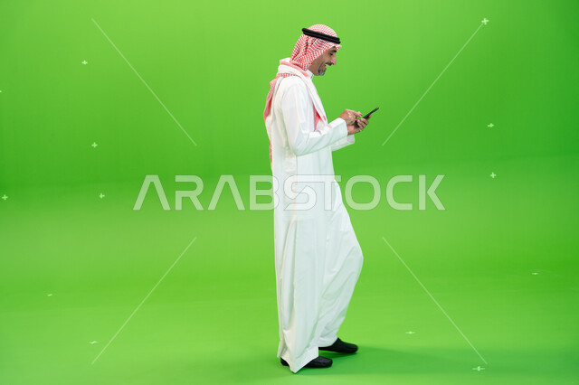 التنقل في المكان أثناء التواصل مع الأقارب والأصدقاء بإيماءات السعادة، إرسال رسائل نصيه عبر الجوال، كروما مقربة لرجل عربي خليجي سعودي يرتدي الزي التقليدي السعودي يقوم بمتابعه أخبار السوشال ميديا عبر الهاتف المحمول، صورة الجسم بالطول الكامل، خلفية خضراء