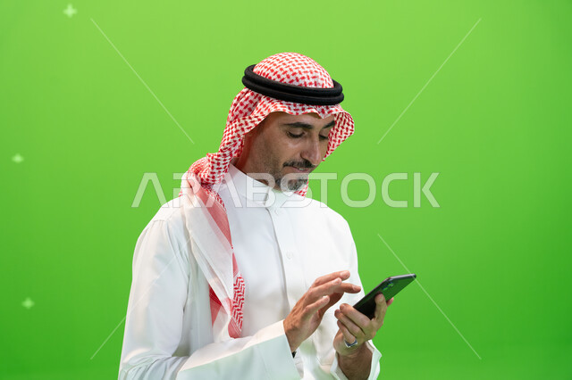 التواصل مع الأقارب والأصدقاء بإيماءات السعادة، إرسال رسائل نصيه عبر الجوال، مفهوم صلة الرحم، كروما مقربة لرجل عربي خليجي سعودي يرتدي الزي التقليدي السعودي يقوم بمتابعه أخبار السوشال ميديا عبر الهاتف المحمول، خلفية خضراء