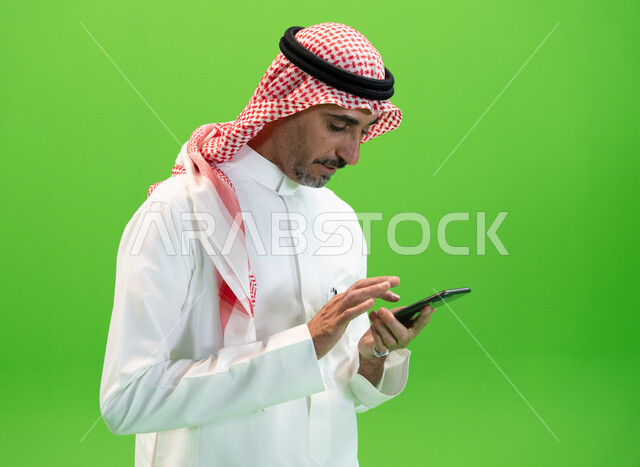 التواصل مع الأقارب والأصدقاء بإيماءات السعادة، إرسال رسائل نصيه عبر الجوال، مفهوم صلة الرحم، كروما مقربة لرجل عربي خليجي سعودي يرتدي الزي التقليدي السعودي يقوم بمتابعه أخبار السوشال ميديا عبر الهاتف المحمول، خلفية خضراء