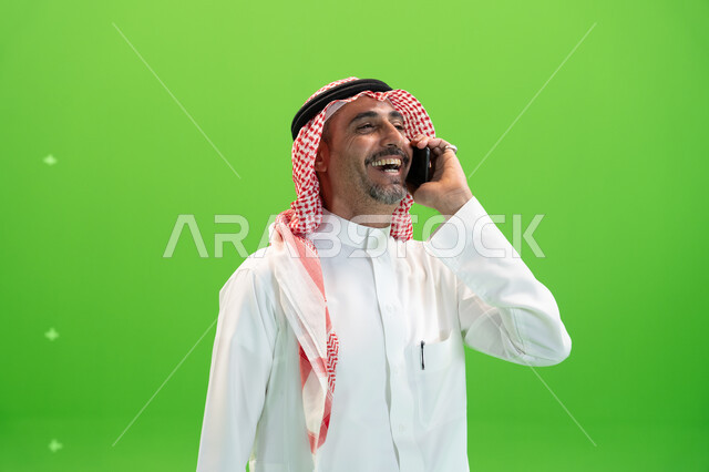  تبادل الاحاديث المسلية والمضحكة، الضحك والتسامر مع الأصدقاء والأقارب بإيماءات السعادة، مفهوم صلة الرحم، كروما مقربة لرجل عربي خليجي سعودي يرتدي الثوب والشماغ السعودي يقوم بإجراء مكالمة صوتيه عبر الهاتف المحمول، خلفية خضراء