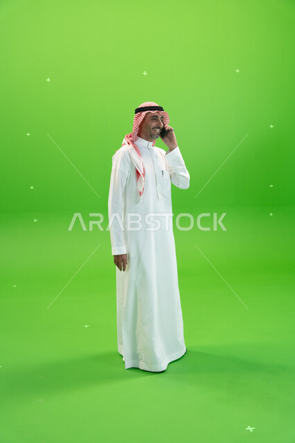 التسامر والضحك الأصدقاء والأقارب بإيماءات السعادة، مفهوم صلة الرحم، الاتفاق على موعد عمل، كروما لرجل عربي خليجي سعودي يرتدي الثوب والشماغ السعودي يقوم بإجراء مكالمة صوتيه عبر الهاتف المحمول، صورة الجسم بالطول الكامل، خلفية خضراء