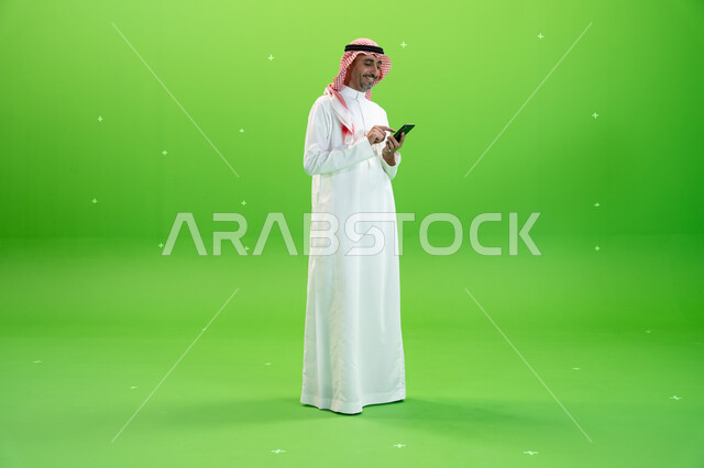إرسال رسائل نصيه عبر الجوال، مفهوم صلة الرحم، كروما لرجل عربي خليجي سعودي يرتدي الثوب والشماغ السعودي يقوم بمتابعه أخبار السوشال ميديا عبر الهاتف المحمول، التواصل مع الأصدقاء والأقارب بإيماءات السعادة، صورة الجسم بالطول الكامل، خلفية خضراء