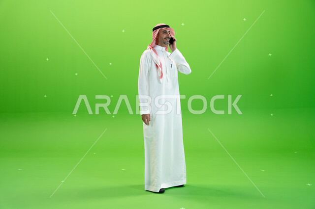 الاتفاق على موعد عمل، كروما لرجل عربي خليجي سعودي يرتدي الثوب والشماغ السعودي يقوم بإجراء مكالمة صوتيه عبر الهاتف المحمول، التواصل مع الأصدقاء والأقارب بإيماءات السعادة، مفهوم صلة الرحم، صورة الجسم بالطول الكامل، خلفية خضراء