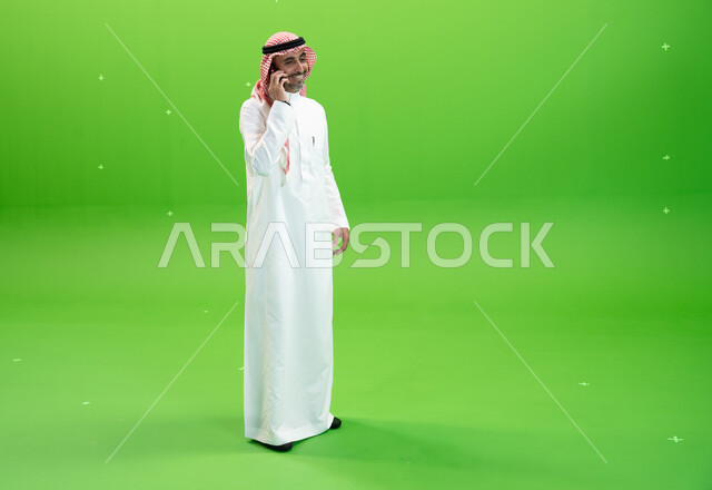 الاتفاق على موعد عمل، كروما لرجل عربي خليجي سعودي يرتدي الثوب والشماغ السعودي يقوم بإجراء مكالمة صوتيه عبر الهاتف المحمول، التواصل مع الأصدقاء والأقارب بإيماءات السعادة، مفهوم صلة الرحم، صورة الجسم بالطول الكامل، خلفية خضراء