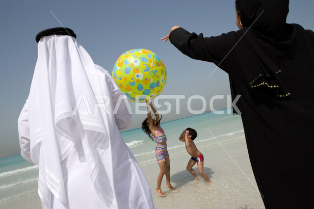 مفهوم الأسرة الإماراتية المترابطة, صورة من الخلف لعائلة عربية سعودية خليجية تقوم باللعب بالكرة المطاطية على شاطئ البحر, قضاء أوقات الإجازة مع الأبناء, السياحة في دول الخليج العربي, مشاركة العائلة الوقت الترفيهي الممتع