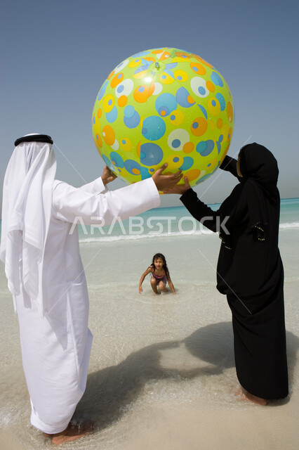 الاستمتاع والسياحة في دول الخليج العربي, منتجعات الشاطئ الإماراتي, صورة من الخلف لأب وأم عربيين خليجيين إماراتيين يلعبان بالكرة المطاطية على شاطئ البحر, مفهوم الأسرة السعيدة, الترابط الأسري وقضاء وقت ممتع مع الأبناء