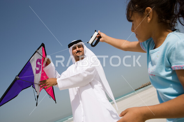 إيماءات السعادة والاستمتاع بقضاء الإجازة الصيفية مع الأبناء , أب عربي خليجي إماراتي يرتدي الكندورة والغترة الإماراتية يقوم باللعب مع ابنته بالطائرة الورقية, منتجعات شاطئية في الامارات , أنشطة ترفيهية مسلية في العطلة