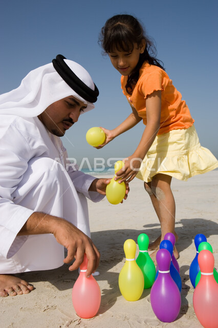The use of plastic bowling pins in play, the concept of parental bonding, enjoying recreational activities with children, an Arab Emirati father wearing a kandora and a ghutra playing with his daughter on the beach, the importance of sharing fun leisure t