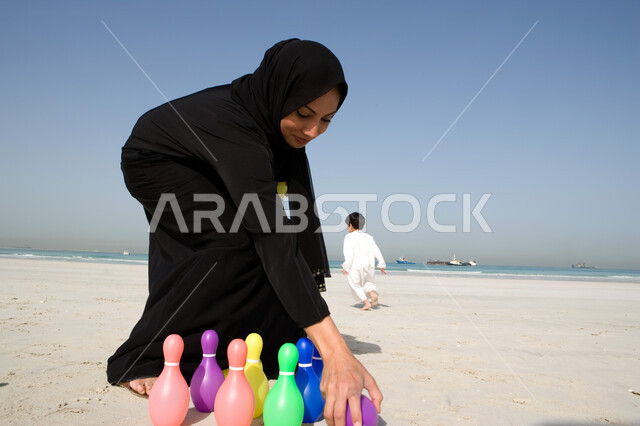القيام بالأنشطة الترفيهيه المختلفة مع الأبناء, استخدام دبابيس البولينج البلاستيكية في اللعب, أم عربية خليجية إماراتية ترتدي العباءة السوداء والحجاب تقوم بترتيب لعبة البولينج, الاستمتاع بقضاء وقت الإجازة الصيفية مع الأسرة