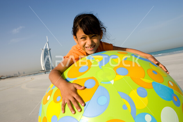 قضاء الإجازة الصيفية على البحر بإيماءات السعادة والفرح, المنتجعات والشواطئ في الإمارات, صورة مقربة لفتاة صغيرة عربية إماراتية خليجية مبتسمة تمسك بيدها الكرة المطاطية الكبيرة على الشاطئ, أنشطة ترفيهية مسلية في العطلة