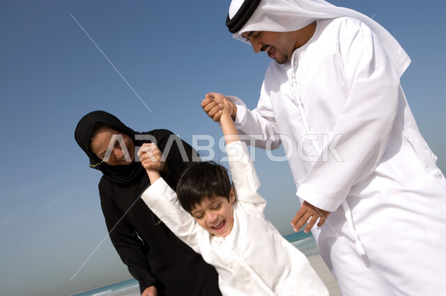 الاستمتاع في منتجعات الشاطئ الإماراتي, قضاء أوقات الإجازة مع الأهل بإيماءات السرور والفرح, أب وأم عربيان خليجيان إماراتيان يمسكان يد ابنهم عاليا على شاطئ البحر, السعادة العائلية برفقة الأبناء