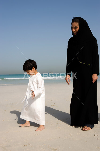 السير مع الأبناء على شاطئ البحر, القيام بالفعاليات الصيفية مع الأبناء, أم عربية خليجية إماراتية ترتدي العباءة السوداء تنظر إلى ابنها وتقوم باللعب معه على الشاطئ, الاستمتاع بقضاء أوقات ممتعه مع الأولاد, منتجعات وشواطئ الإمارات