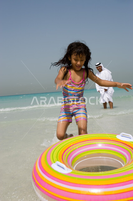 أنشطة ترفيهية مسلية في العطلة, فتاة صغيرة تقوم باللعب بعوامة السباحة, إيماءات السعادة والاستمتاع بقضاء الإجازة الصيفية مع الأبناء , أب عربي خليجي إماراتي يرتدي الكندورة والغترة الإماراتية يقوم باللعب مع ابنته على الشاطئ, منتجعات شاطئية في الامارات