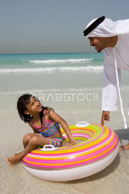 Gestures of happiness and enjoying the summer vacation with the children, entertaining activities during the holiday, a young girl sitting on a swimming buoy, an Emirati Arab father wearing an Emirati kandora and ghutra playing with his daughter on the be