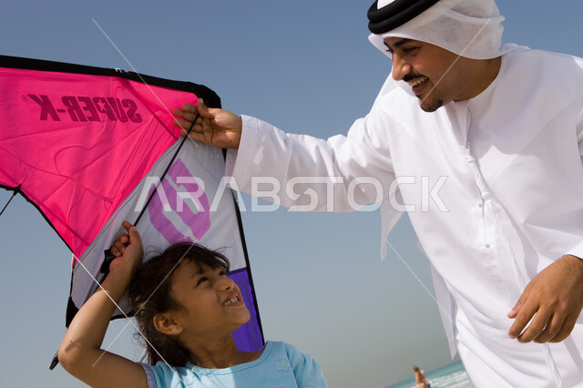 Paying attention to children's hobbies, gestures of happiness and enjoying spending the summer vacation with the family, an Arab Emirati father wearing an Emirati kandora and ghutra playing with his daughter in a kite, beach resorts in the Emirates, fun l