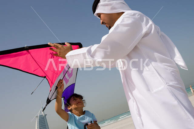 أنشطة ترفيهية ممتعه في العطلة, الاهتمام بهوايات الأبناء, إيماءات السعادة والاستمتاع بقضاء الإجازة الصيفية مع العائلة, أب عربي خليجي إماراتي يرتدي الكندورة والغترة الإماراتية يقوم باللعب مع ابنته بالطائرة الورقية, منتجعات شاطئية في الامارات