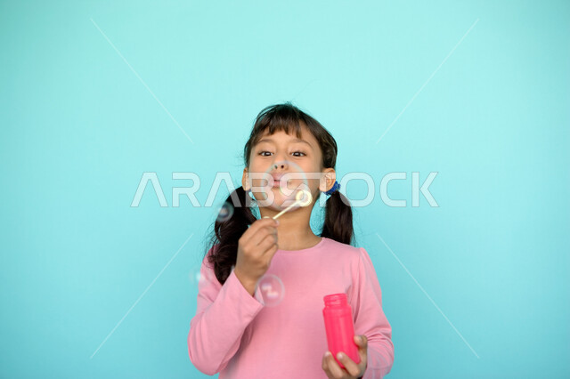 قضاء الأوقات بالأنشطة الترفيهية الممتعه والمسلية, بورتريه مقرب لفتاة عربية خليجية إماراتية تقوم بنفخ البالونات الهوائية, اللعب بالفقاعات المائية المتطايرة والمتناثرة, تنمية مهارات الأطفال الحركية, الاستمتاع بصنع فقاعة الصابون في الهواء, خلفية ملونة