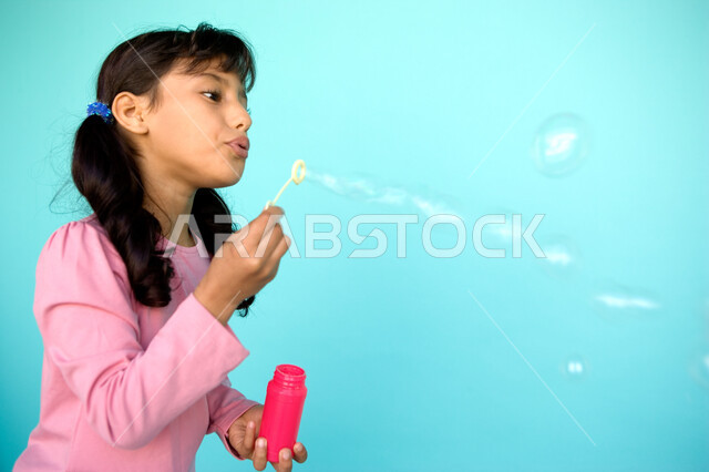 اللعب بالفقاعات المائية المتطايرة والمتناثرة, قضاء الأوقات بالأنشطة الترفيهية الممتعه والمسلية, بورتريه مقرب لفتاة عربية خليجية إماراتية تقوم بنفخ البالونات الهوائية, تنمية مهارات الأطفال الحركية, الاستمتاع بصنع فقاعة الصابون في الهواء, خلفية ملونة