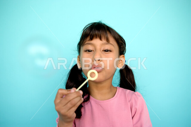 الأنشطة الترفيهية الممتعه والمسلية, بورتريه مقرب لفتاة عربية خليجية إماراتية تقوم بنفخ البالونات الهوائية, اللعب بالفقاعات المائية المتطايرة والمتناثرة, تنمية مهارات الأطفال الحركية, الاستمتاع بصنع فقاعة الصابون في الهواء, خلفية ملونة