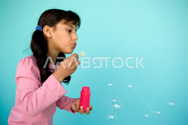 المتعة والتسلية بقضاء الأنشطة الترفيهية, بورتريه مقرب لفتاة عربية خليجية إماراتية تقوم بنفخ البالونات الهوائية, اللعب بالفقاعات المائية المتطايرة والمتناثرة, تنمية مهارات الأطفال الحركية, الاستمتاع بصنع فقاعة الصابون في الهواء, خلفية ملونة