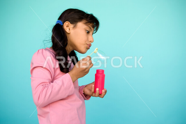 التسلية والمتعه بقضاء الأنشطة الترفيهية, بورتريه مقرب لفتاة عربية خليجية إماراتية تقوم بنفخ البالونات الهوائية, اللعب بالفقاعات المائية المتطايرة والمتناثرة, تنمية مهارات الأطفال الحركية, الاستمتاع بصنع فقاعة الصابون في الهواء, خلفية ملونة