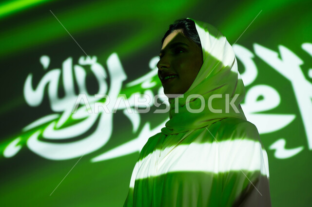 انغماس الجسم في ضوء العلم، مفهوم حب الوطن والافتخار والاعتزاز به، إحياء ذكرى اليوم الوطني السعودي 23 سبتمبر، الاحتفال  بيوم العلم 11 مارس والعيد الوطني، أعياد ومناسبات وطنية، صورة مقربة من الجانب لامرأة عربية خليجية سعودية محجبة تنظر إلى شيء ما