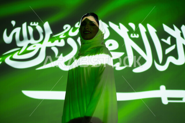 الوقوف بثقة وفخر والنظر إلى الكاميرا، مفهوم حب الوطن والانتماء إليه، ذكرى اليوم الوطني السعودي 23 سبتمبر، الاحتفال في يوم العلم 11 مارس والعيد الوطني، أعياد ومناسبات وطنية ، انغماس جسد امرأة عربية خليجية سعودية محجبة مبتسمة في ضوء العلم