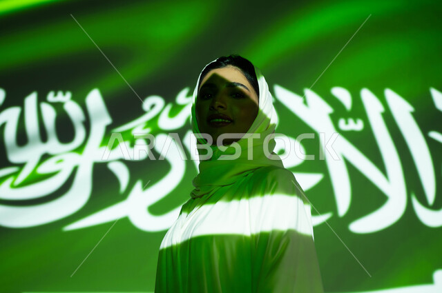 الوقوف بثقة والنظر إلى الكاميرا، اندماج الجسم مع ضوء العلم، الافتخار بالهوية الوطنية السعودية، إحياء ذكرى اليوم الوطني السعودي 23 سبتمبر، يوم العلم 11 مارس، أعياد ومناسبات وطنية ، صورة مقربة لامرأة عربية خليجية محجبة ترتدي العباءة، الاحتفال في العيد الوطني