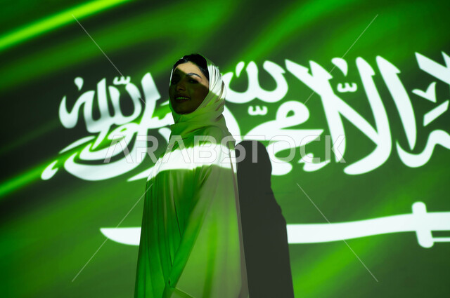 الوقوف بثقة وفخر والنظر إلى الكاميرا، مفهوم حب الوطن والانتماء إليه، إحياء ذكرى اليوم الوطني السعودي 23 سبتمبر، يوم العلم 11 مارس، أعياد ومناسبات وطنية ، انغماس جسد امرأة عربية خليجية سعودية محجبة مبتسمة في ضوء العلم، الاحتفال في العيد الوطني .