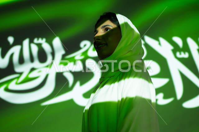 الافتخار والاعتزاز بالتاريخ والهوية الوطنية، الاحتفال بالعيد الوطني، ذكرى اليوم الوطني السعودي 23 سبتمبر، يوم العلم السعودي 11 مارس، أعياد ومناسبات شعبيه، انغماس الجسم في ضوء العلم، صورة مقربة من الجانب لامرأة عربية خليجية سعودية محجبة تنظر إلى شيء ما