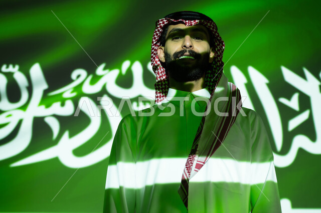 الفخر والتباهي بالهوية الوطنية السعودية، الوقوف بثقة و فخر والنظر إلى الكاميرا، انعكاس ضوء العلم على الوجه، صورة مقربة لرجل عربي خليجي سعودي يرتدي الزي التقليدي, إحياء ذكرى اليوم الوطني السعودي 23 سبتمبر، الاحتفال بيوم العلم 11 مارس و العيد الوطني