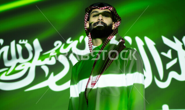 الفخر والتباهي بالهوية الوطنية السعودية، الوقوف بثقة و فخر والنظر إلى الكاميرا، انعكاس ضوء العلم على الوجه، صورة مقربة لرجل عربي خليجي سعودي يرتدي الزي التقليدي, إحياء ذكرى اليوم الوطني السعودي 23 سبتمبر، الاحتفال بيوم العلم 11 مارس و العيد الوطني