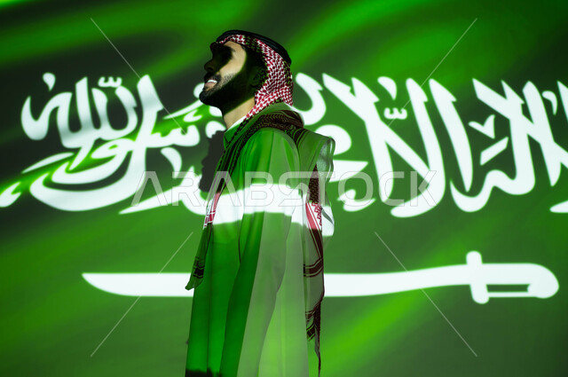 Pride in the Saudi national identity, immersion of the body in the light of the flag, a side image of a Gulf Arab man wearing traditional Saudi dress, looking at something, commemorating the Saudi National Day 93, the Saudi Flag Day on March 11, the Natio