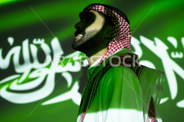 إظهار مشاعر الحب والولاء إلى السعودية، انغماس ضوء العلم مع الوجه، الاحتفال بيوم العلم 11مارس والعيد الوطني، أعياد ومناسبات وطنية ،صورة مقربة من الجانب لرجل عربي خليجي سعودي مبتسم يرتدي الزي التقليدي ينظر إلى شيء ما, ذكرى اليوم الوطني السعودي 23 سبتمبر .