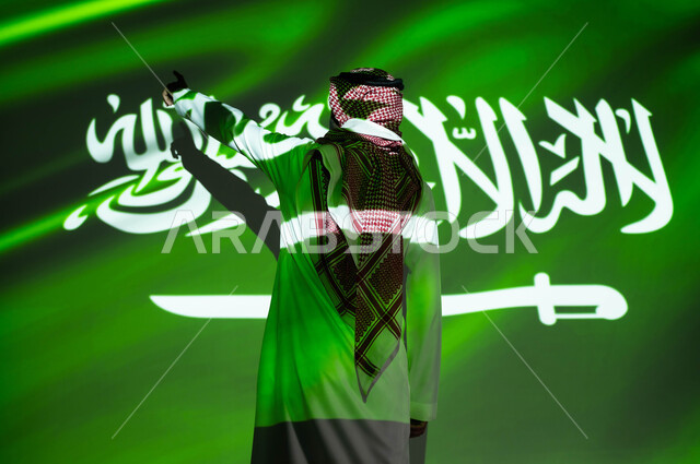 الاعتزاز والفخر بالهوية السعودية، إحياء ذكرى اليوم الوطني السعودي 23 سبتمبر، يوم العلم 11 مارس، أعياد ومناسبات وطنية ، اندماج الجسم في ضوء العلم، صورة من الخلف لرجل عربي خليجي سعودي يرتدي الزي التقليدي يشير إلى شيء ما يحتفل في العيد الوطني