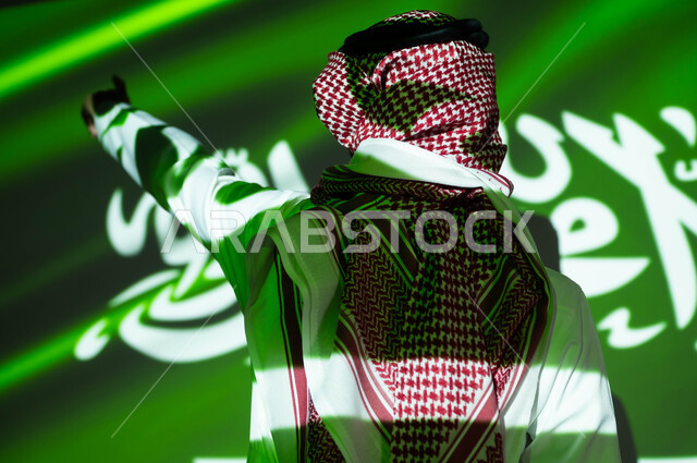 الاحتفال في العيد الوطني ،الاعتزاز والفخر بالهوية السعودية، إحياء ذكرى اليوم الوطني السعودي 23 سبتمبر، يوم العلم 11 مارس، أعياد ومناسبات وطنية ، اندماج الجسم في ضوء العلم، صورة مقربة من الخلف لرجل عربي خليجي سعودي يرتدي الزي التقليدي يشير إلى شيء ما