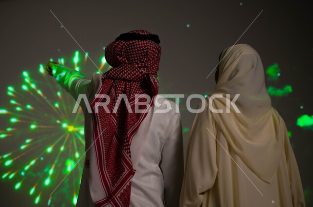 الاستمتاع بالمفرقعات في الأعياد الوطنية، إظهار الولاء و التفاني للبلاد، رجل عربي خليجي سعودي يرتدي الزي التقليدي يشير بيده إلى الألعاب النارية، صورة من الخلف لزوجين خليجيين سعوديين يحتفلان بذكرى اليوم الوطني السعودي في23 سبتمبر والعيد الوطني
