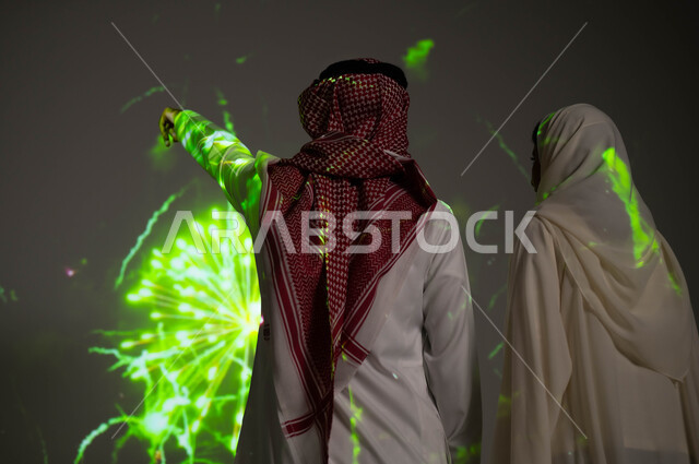 الاستمتاع بالألعاب النارية في الأعياد الوطنية، إظهار الولاء و التفاني للبلاد، رجل عربي خليجي سعودي يرتدي الزي التقليدي يشير بيده إلى المفرقعات، صورة من الخلف لزوجين خليجيين سعوديين يحتفلان بذكرى اليوم الوطني السعودي في23 سبتمبر والعيد الوطني