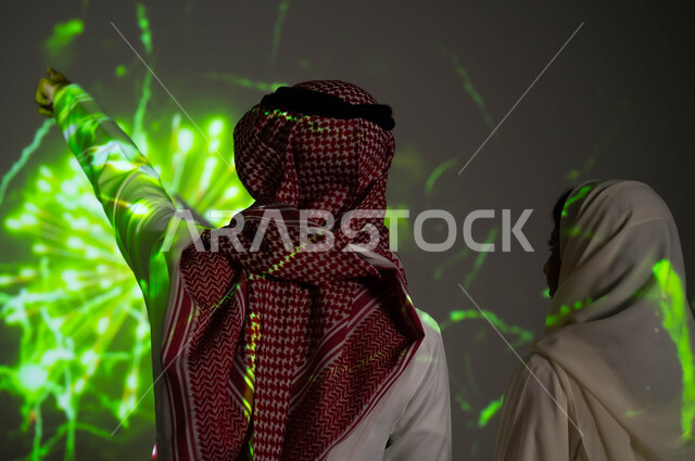 إظهار التفاني للبلاد، الاستمتاع بالألعاب النارية في الأعياد الوطنية، رجل عربي خليجي سعودي يرتدي الزي التقليدي يشير بيده إلى المفرقعات، صورة مقربة من الخلف لزوجين خليجيين سعوديين يحتفلان بذكرى اليوم الوطني السعودي في23 سبتمبر والعيد الوطني